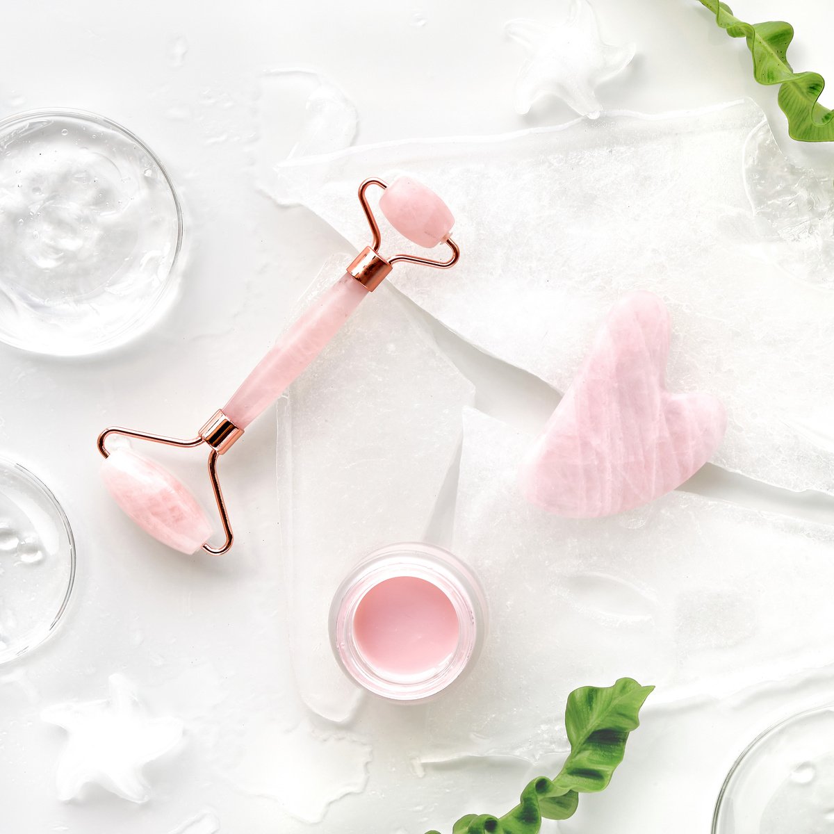 Pink Quarts Face Roller, Gua Sha Stone, Pink Cream in Glass Jar. off White Flat Lay in Green and Pink with Ice and Exotic Fern Leaves. Beauty Facial Massage with Natural Cosmetic Ingredients.